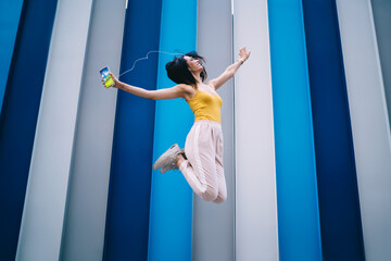 Excited hipster girl in headphones rejoicing during positive leisure time with energetic music podcast, joyful female millennial with smartphone listening funny media playlist dancing and jump