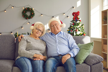 Happy senior couple in Santa hats looking at camera while sitting on couch at home