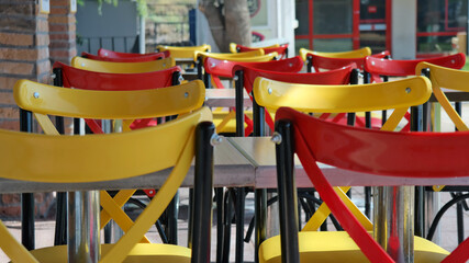 Fototapeta na wymiar Tables and metal chairs with vivid red and yellow colors in harmony with green trees and blue sky in an open air restaurant at lunch time in a university campus, empty because of the Covid-19 pandemic