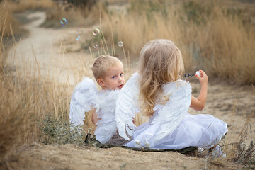 Two little angels boy and girl sitting back on a hill plaing soap bubbles. Girl blowing soap balls. 