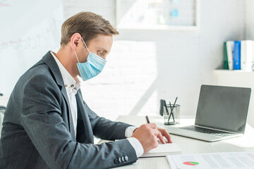 Side view of businessman in medical mask, writing in blank notebook, while sitting at workplace on...