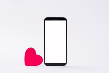 A pink heart stands beside a smartphone with a blank white display. Items on a white background. Concept of declaration of love with through messages.