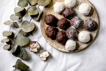 Raw vegan homemade coconut chocolate candy balls whole and broken with coconut flakes in ceramic plate over white textile background with eucalyptus branches. Flat lay, space