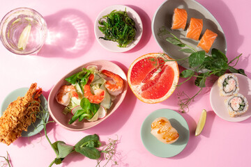 Sushi set flat lay on pink background. Creative layout of asian food. Maki roll with salmon, California roll, scallop sashimi nigiri, wakame, fried shrimps, prawn salad, fruits. Bright close up