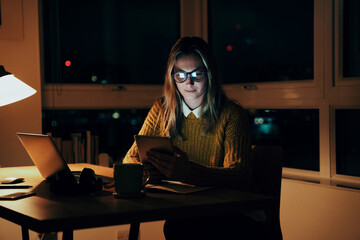 Caucasian business woman working at office desk busy with business report reading emails on digital tablet sitting in apartment 