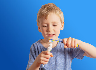 a boy with white hair holds a brush in his hands and squeezes the paste out of the tube onto the brush. blue background.