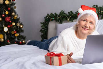 Obraz na płótnie Canvas Christmas online greetings. happy senior women in Santa hat at home in bed with laptop.
