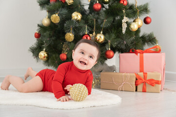 baby girl 6 month in red bodysuit under Christmas tree with gift boxes presents. Christmas lights.