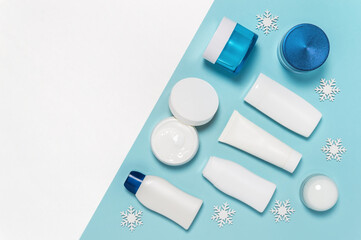 Set of winter skin care cosmetic products on white and blue background with snowflakes. Cream jar, tube, body lotion, milk and cleanser bottle package mockup. Flat lay, copy space
