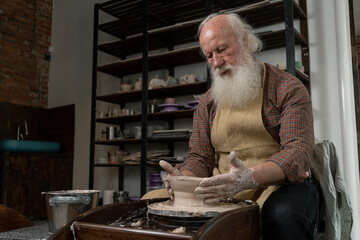 Old male potter. High angle of craftsman working on pottery wheel while sculpting from clay pot in workshop. Focus on arms. Concept of ceramic art