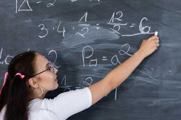 A teenage girl is solving a math problem at school by writing on a school blackboard with white chalk. Her hair is pinned with a pink ribbon in two ponytails.