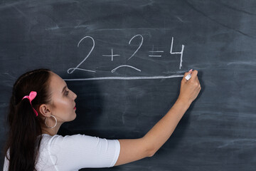 Teenager chalks a math equation on a dark gray school blackboard. Her hair is pinned with a pink ribbon in two ponytails.