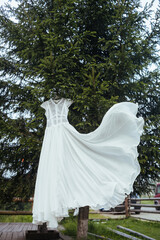 Wedding white lace long dress hangs on a hanger on the green tree. Stylish clothes for wedding ceremony outdoors. Morning and preparation of the bride.