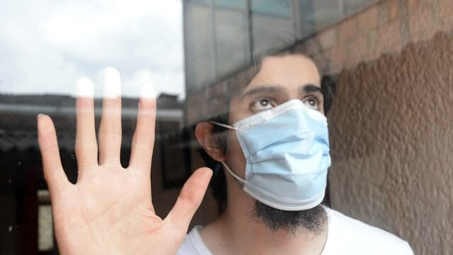 Latino male model in white shirt and light blue medical mask looking through the window with a cloudy sky and buildings reflexed. Scene of new normality during the covid 19 pandemic