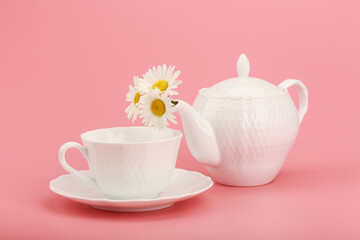 Creative concept of a healthy lifestyle and herbal tea. White tea pot with camomile flowers and a cup on bright pink background