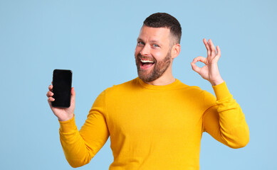 funny cheerful man in a yellow sweater with phone makes a gesture of approval on blue background.