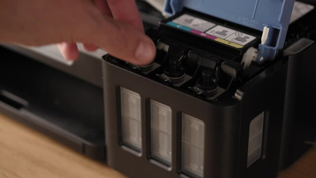 Video instruction, technology, media, office, stationery concept - close-up of male hands preparing new inkjet printer for launch. step 9: pouring ink - grab and remove black rubber tank cap of tank