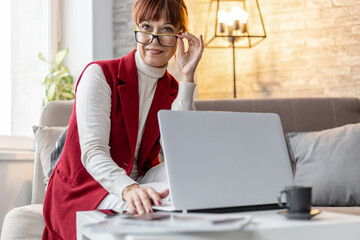 an adult woman with glasses sits on a sofa and works at a laptop at home