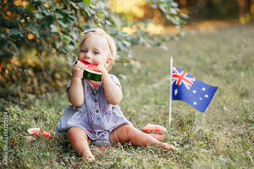 Cute adorable Caucasian baby girl eating ripe watermelon with Australian flag on ground. Funny child kid sitting in park with fresh fruit celebrating Australia Day holiday outdoors.