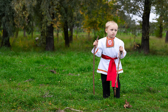 a boy in a Russian national costume stands in the grass in his hands two wooden sticks