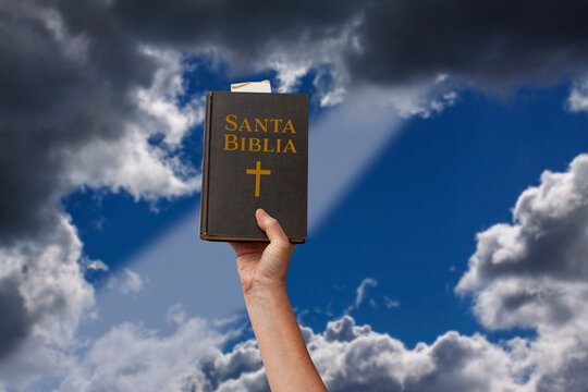 Arm raised into the air a hand reaching up and holding the Holy Bible in Spanish Santa Biblia. Ray of light coming through dramatic blurred clouds . Symbolic posture of prayer, devotion and worship.