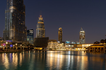 Night view of illuminated skyscrapers and waterfront at downtown Dubai, United Arab Emirates. Golden tower blocks and blue-glass buildings rise above the Burj Lake, reflecting on calm water under a cl