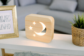 Close up cropped image of decorative wooden night lamp with moon and stars picture, on light table with flower pot and photo frame. Cozy living room interior with gray sofa on the background