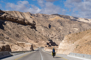 Sunlit mountain pass road winds through arid hills in Oman’s Al Hajar Mountains. A lone motorcyclist rides along the asphalt highway carved between rocky slopes under a clear blue sky in January 2020.