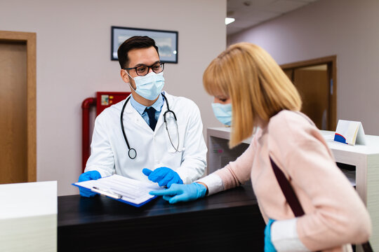 Young Male Practitioner With Face Protective Mask And Gloves Working At Clinic Reception Desk And Talking With Patient Or Visitor.