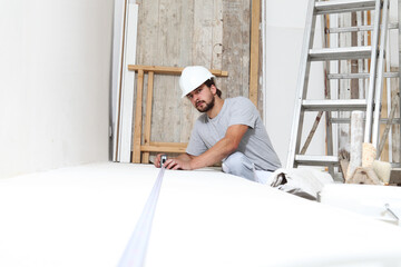 construction worker plasterer man measuring wall with measure tape in building site of home renovation with tools and building materials on the floor