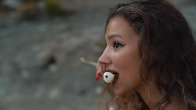 Pretty young woman with long dark brown hair holds wooden stick tastes and enjoys hot and stringy marshmallow piece roasted on fire close view slow motion.