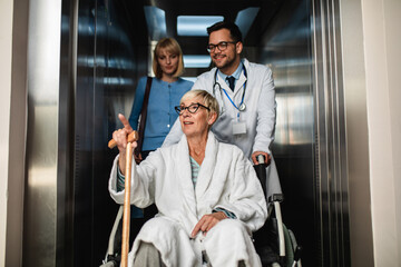 Young male practitioner talking with middle age woman and her sick mother which sitting in disability chair. They are standing in hospital or nursing center elevator.