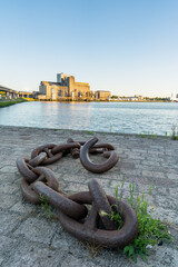 Close-up of a heavy iron mooring chain leading the eye toward a historic waterfront building on the Nieuwe Maas river in Rotterdam, Netherlands. Weathered concrete and rippling water create a gritty p
