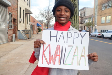 Black kid holding Happy Kwanzaa sign outdoors on city street