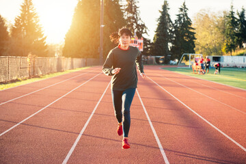 Fototapeta na wymiar Asian woman running on the track in the winter. Sun flare suggests sunrise or sunset.