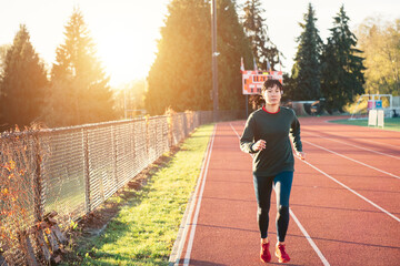 Asian woman running on the track in the winter. Sun flare suggests sunrise or sunset.