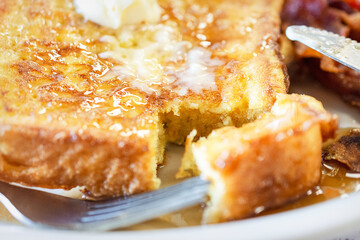 French Toast on a white plate with bacon blurred in the background.  Daub of butter melts ontop with generous amount of maple syrup.  Photo shot on location at diner.