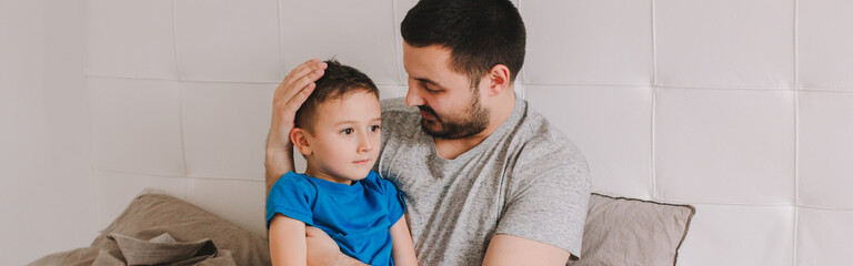 Caucasian father talking to boy son. Man parent hugs kid child on bed in bedroom at home. Authentic...