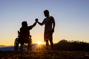 Fototapeta na wymiar Silhouette of joyful disabled man in wheelchair raised hands with friend at sunset