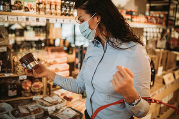 Beautiful young and elegant woman with face protective mask and gloves buying healthy food and drink in a modern supermarket or grocery store. Pandemic or epidemic lifestyle and consumerism concept.