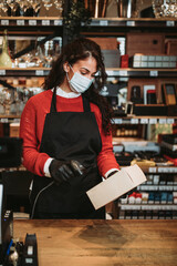 Fototapeta na wymiar Beautiful and positive female cashier with face protective mask working on cash register in a modern supermarket or grocery store. Pandemic or epidemic lifestyle and consumerism concept.