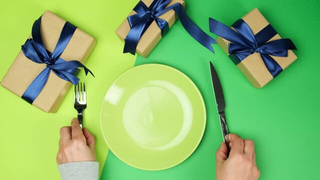 empty round ceramic green plate and female hands hold a fork with a knife on a green background with wrapped gift boxes, top view