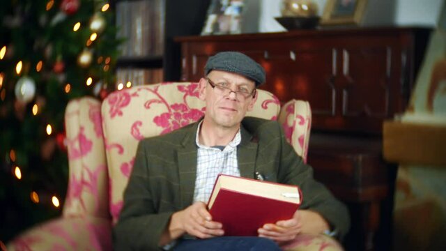 A senior man closes a book after reading a Christmas story. The man is wearing a glasses, a flat cap and tweed jacket. There is a Christmas tree and lights behind him.