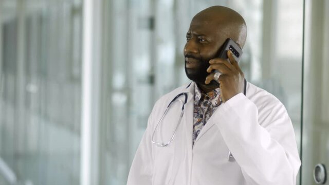 An African Male Doctor Received An Upsetting Phone Call. The Man Is Upset And Sad At Receiving The New As He Talks On The Phone In The Hospital Corridor. He Is Wearing A Lab Coat And Stethoscope.