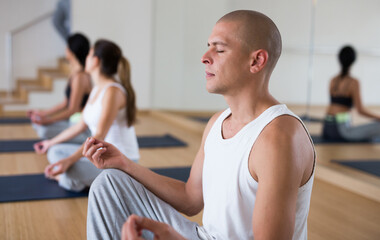 Young man sitting in lotus position with closed eyes practicing meditation at group yoga class
