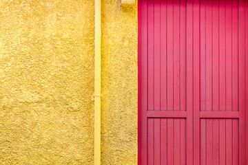 A red wooden door on a yellow wall. An yellow water pipe along the wall.