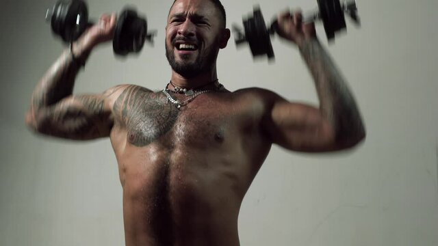 Muscular guy doing exercises with dumbbell. Excited sporty man working out. Muscular naked torso on gray background. Strength and motivation.