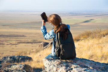 A girl with a backpack sits on a rock above the valley and takes a photo on a smartphone camera