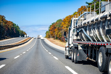 Big rig day cab white semi truck transporting cargo in tank semi trailer for chemicals and other liquids running on the wide straight highway road with autumn trees on the side