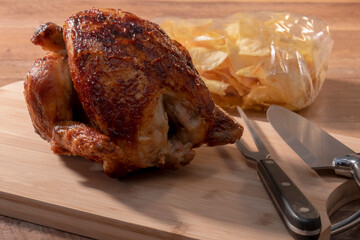 Select focus of a spit-roasted chicken on a wooden board with an out of focus bag of potato chips